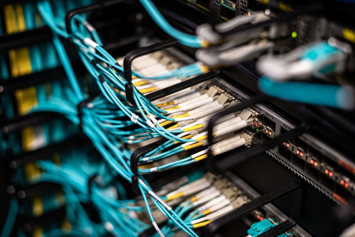 Close-up of a data center switch with dense bundles of aqua fiber optic patch cables plugged into rows of white and yellow LC connectors, routed through black cable management arms with small orange port lights on the right