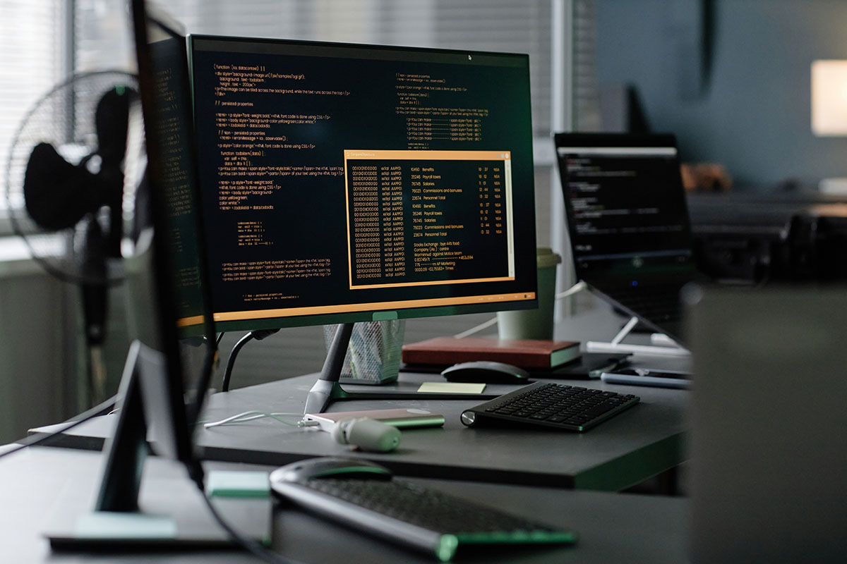 A dark office workstation with a monitor showing lines of code and a data window, a keyboard and a second screen blurred in the background