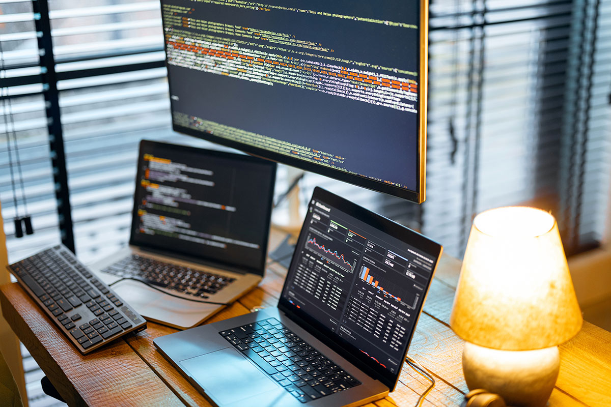 A wooden desk by window blinds with a large monitor and a laptop showing lines of source code, a second laptop showing a dark analytics dashboard, a black keyboard and a lit table lamp