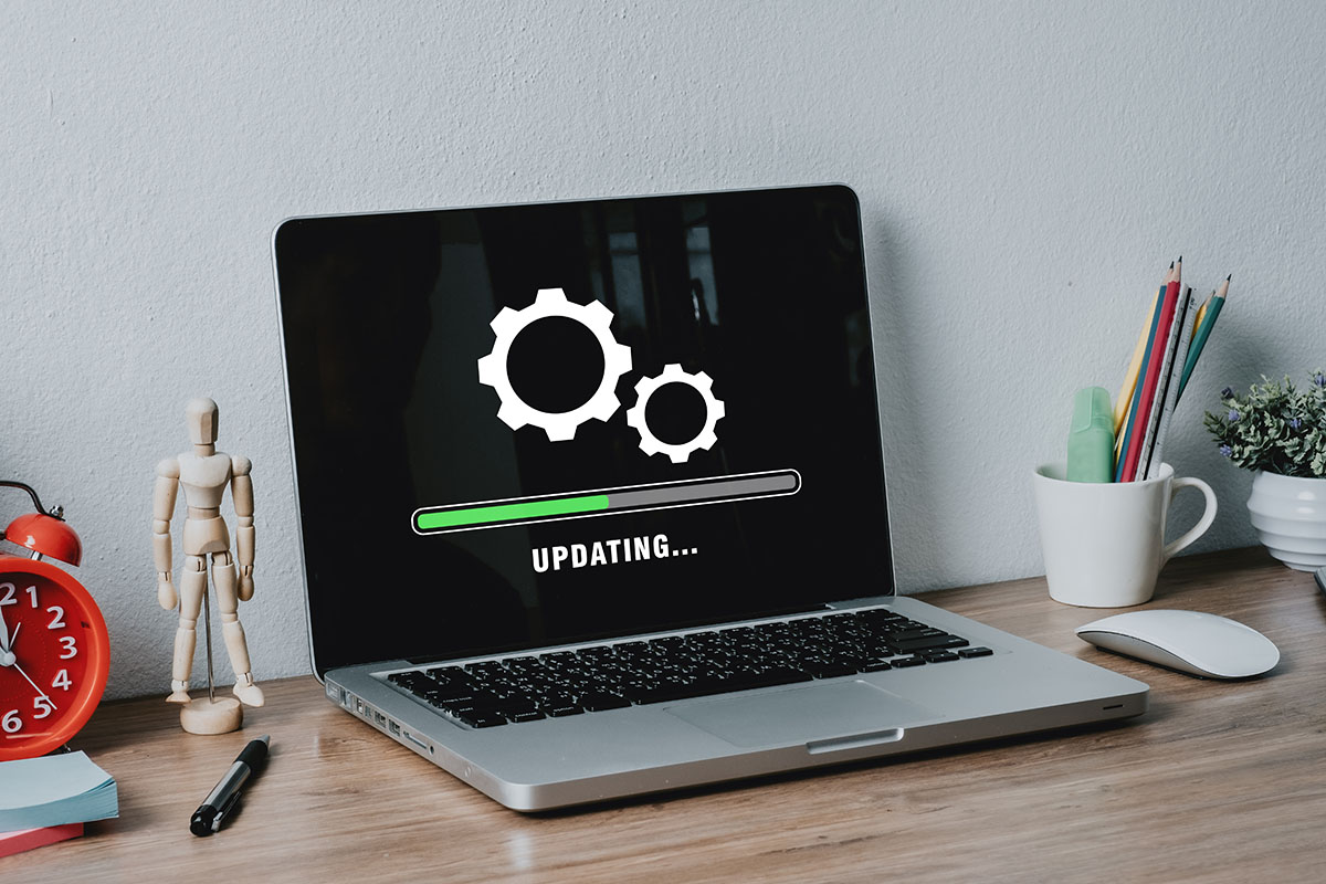 A silver laptop on a wooden desk displaying an UPDATING progress screen, next to a red alarm clock and a wooden artist's mannequin