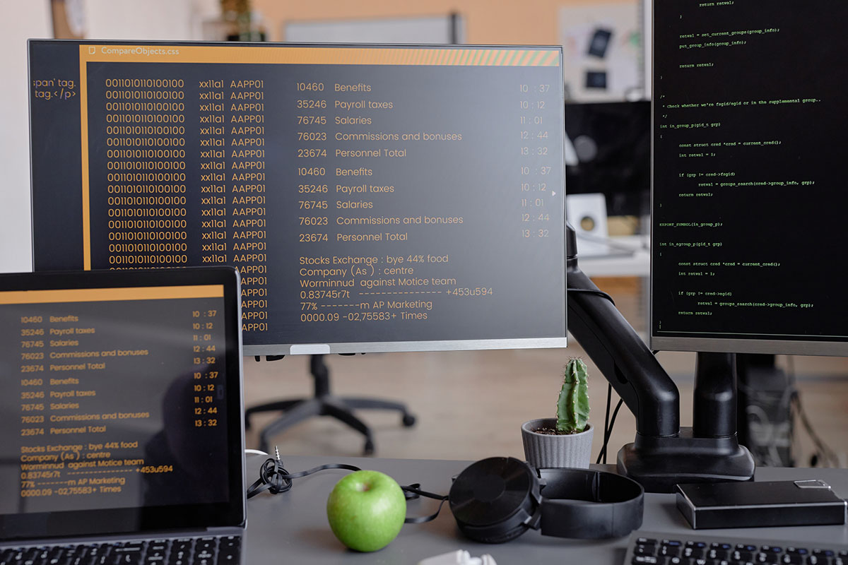 An empty developer desk with a laptop and two monitors showing code and data listings, a monitor arm, headphones, a small cactus and a green apple