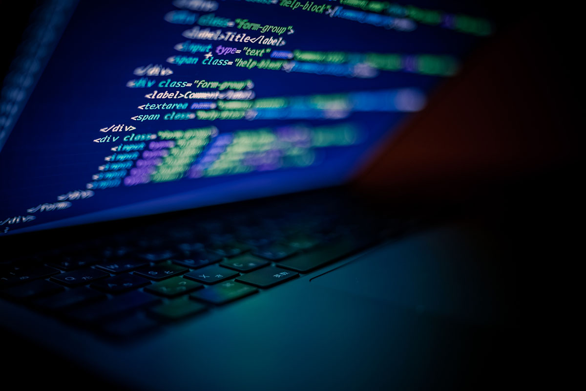 Low-angle close-up of a laptop in a dark room, its screen showing lines of HTML code in blue and green above a dimly lit keyboard