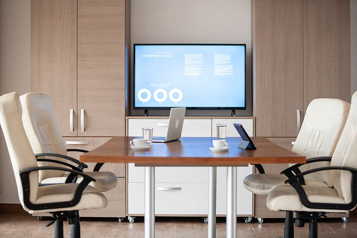 An empty meeting room with white leather chairs around a wooden table, a laptop, tablet and coffee cups, and a wall screen showing a company data presentation