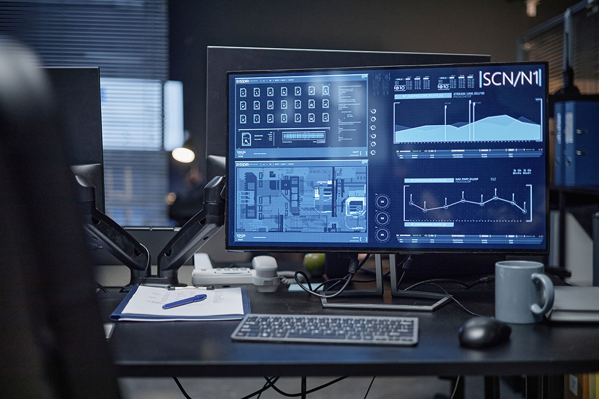 A darkened office desk with dual monitors displaying a security operations dashboard of file counts, access maps, and data flow charts, no people in frame
