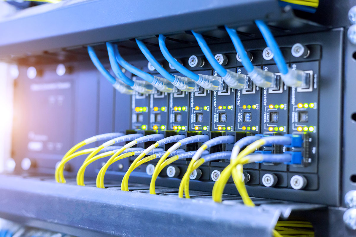 Close-up of a network rack with rows of media converter modules, blue ethernet cables plugged in along the top and yellow fiber patch cords along the bottom, green and amber status lights glowing