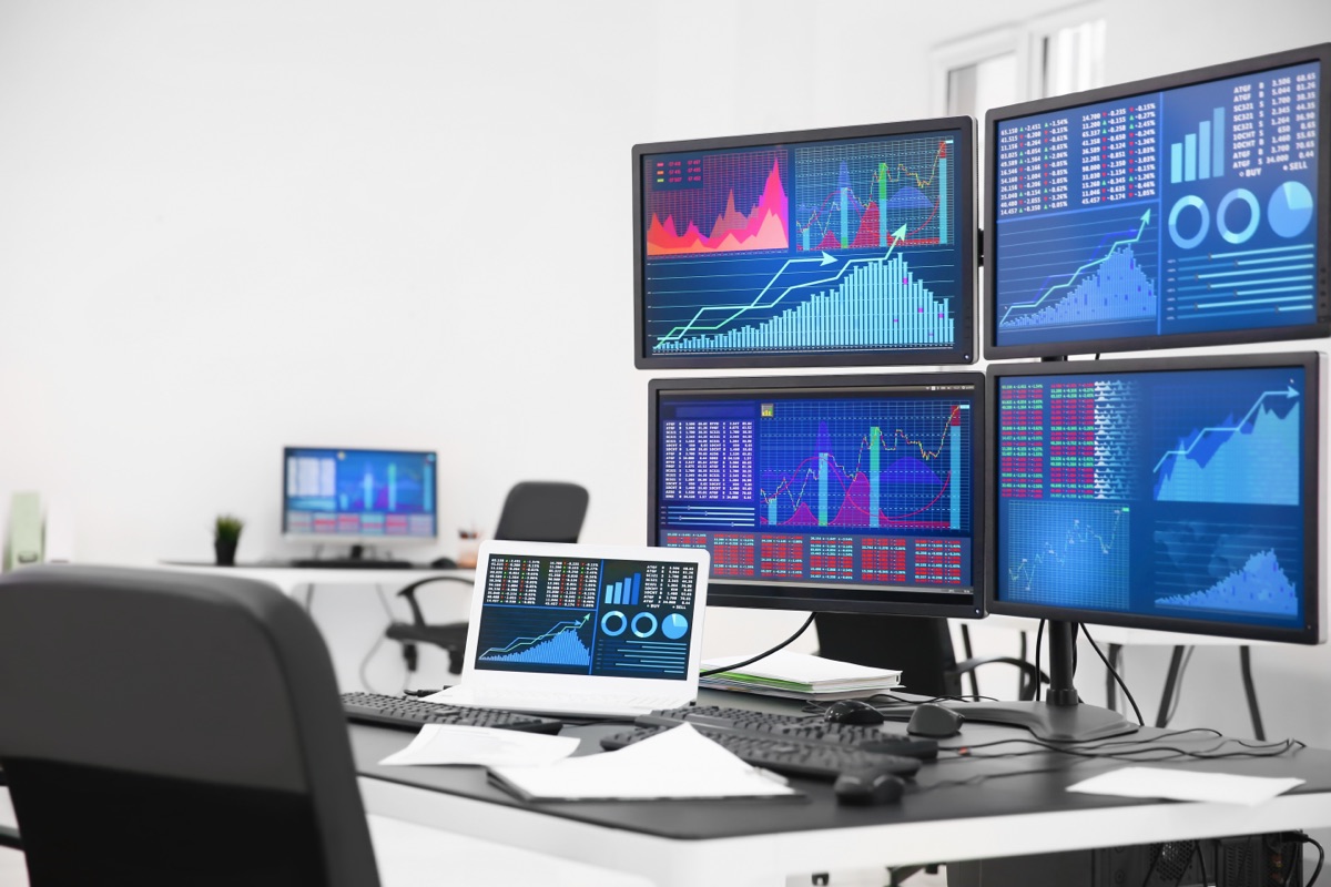 A trading desk in a bright, empty office with four monitors and a laptop showing market charts and price tables