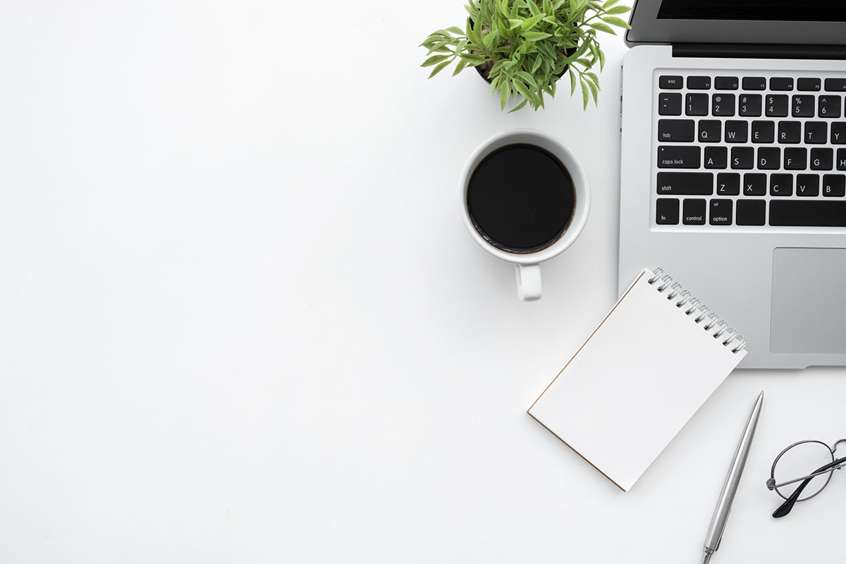 Overhead view of an aluminium laptop, notebook and coffee on a white office desk