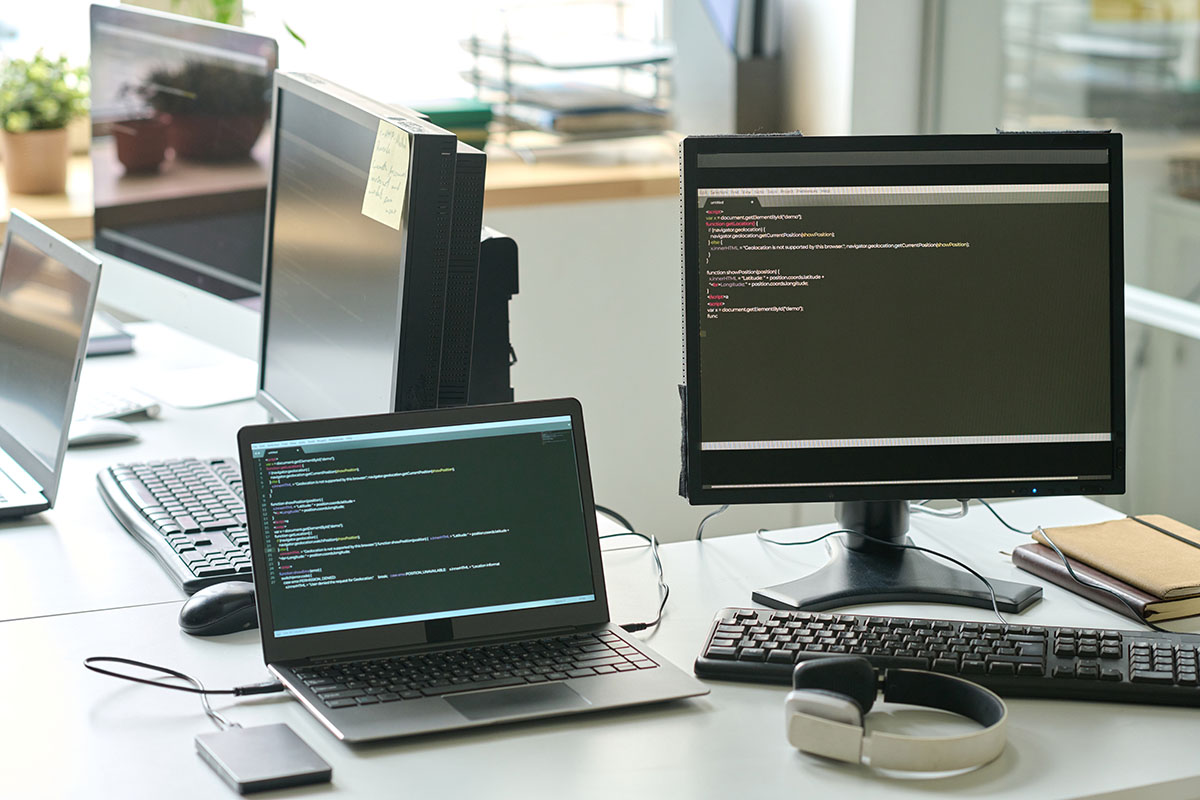 An empty developer workstation on a white office desk with a laptop and a desktop monitor both showing code in a dark editor, alongside keyboards, a mouse, headphones, an external drive and notebooks