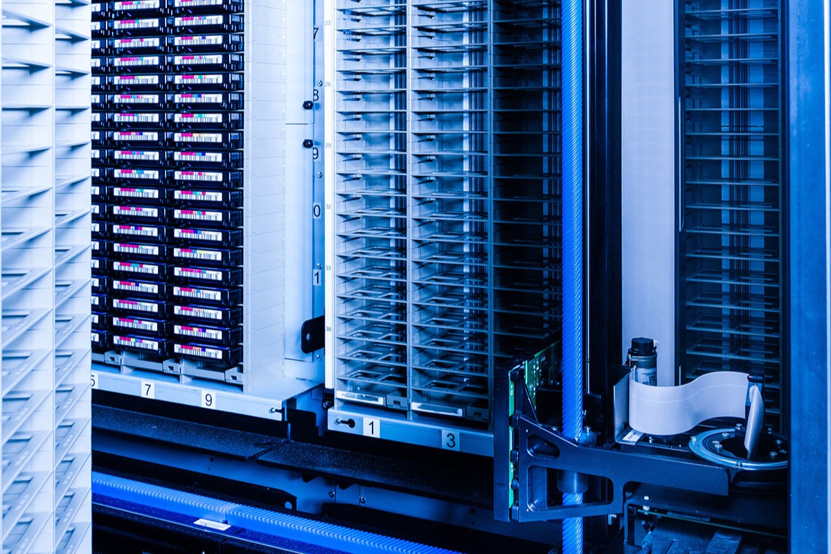Close-up of an automated tape library in a data centre, with rows of labelled backup cartridges in storage slots, empty slot racks and a robotic loader arm lit in blue