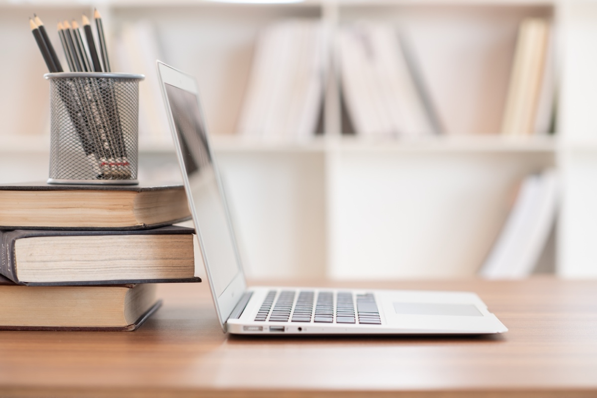 A laptop open beside a stack of books and a pot of pencils in front of a bookshelf