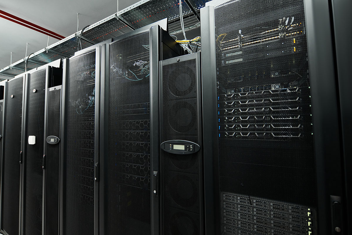 A row of black server cabinets with perforated doors in a data centre, cable trays running along the ceiling above them